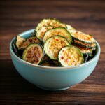 Baked zucchini chips arranged on a tray, golden and crispy, with a sprinkle of seasoning and fresh herbs.